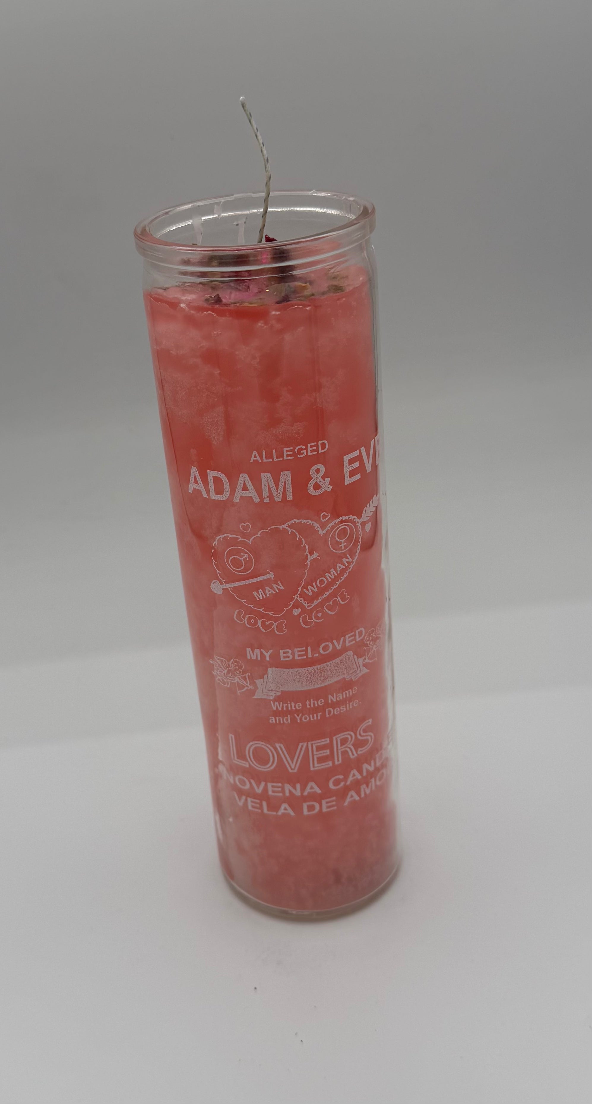 A pink devotional candle in a clear glass jar with a wick, featuring white printed text and heart designs on a neutral gray background.