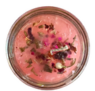 A close-up top-down view of a pink candle in clear glass, containing  dried flower petals, and small pink and green botanical pieces.